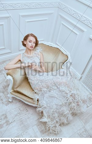 Portrait of a beautiful chic woman posing in a luxury white dress in vintage armchair. Evening makeup and hairstyle of the 20s. 