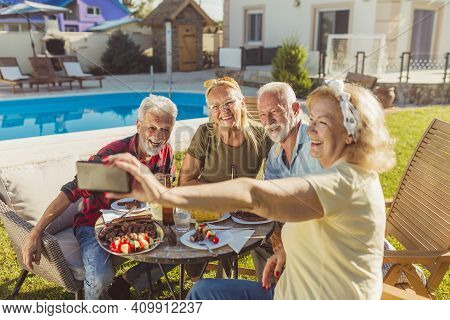 Group Of Elderly Friends Taking A Selfie While Having Lunch In The Backyard By The Pool, Gathered Ar