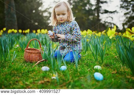 Three Year Old Girl Playing Egg Hunt On Easter. Child Sitting On The Grass With Many Narcissi And Ga