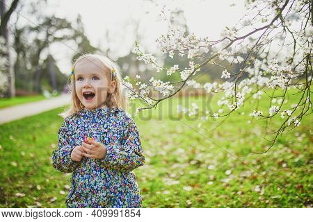 Adorable Little Girl Enjoying Nice And Sunny Spring Day Near Apple Tree In Full Bloom. Outdoor Sprin