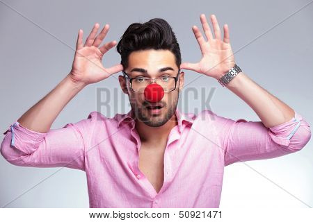 young fashion man with a red nose acting crazy, holding his palms at his head like antlers while looking at the camera. on a light background