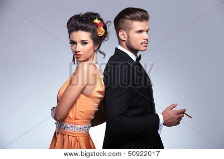 young fashion couple standing back to back while the man is holding a cigarette in his hand. on gray background