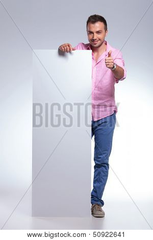 full length photo of a young casual man holding a black pannel and showing the thumb up gesture at the camera with a smile on his face. on gray background