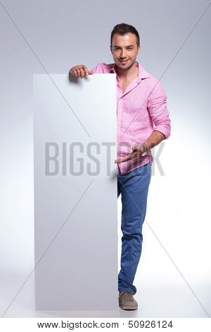 full length photo of a young casual man presenting a blank pannel while smiling for the camera. on gray background