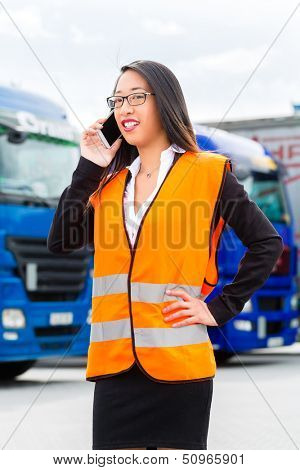 Logistics - female Asian forwarder or supervisor with mobile phone, in front of trucks and trailers, on transshipment point
