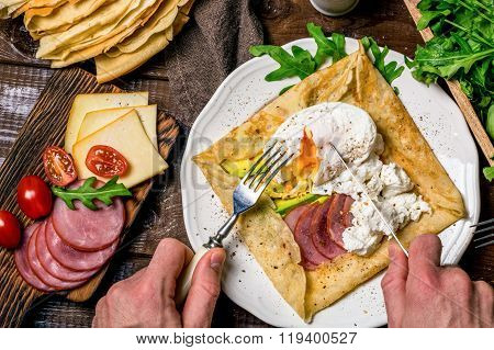 Man eating crepe galette with meat, avocado, soft white cheese and poached egg on white plate. Sliced yellow cheese, pastrami, cherry tomatoes, green salad and stack of crepes on side. Top view