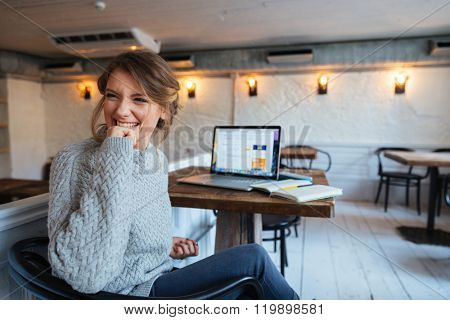 Cheerful woman sitting at the table with laptop computer in cafe and looking away