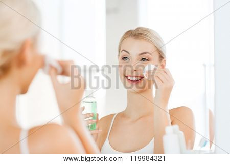 beauty, skin care and people concept - smiling young woman applying lotion to cotton disc for washing her face at bathroom