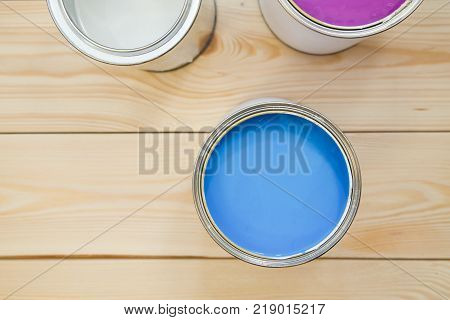 Painting a house. Tin cans of blue, white and fuchsia oil paint on a light uncolored wooden background. Close up. Top view. Space for your text or pruduct display.
