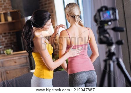 Massage technique. Positive nice young woman standing in front of her friend and looking at her back while presenting massage techniques