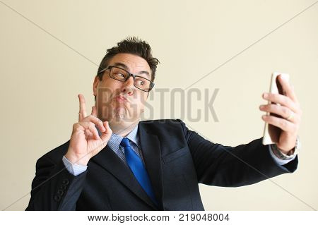 Funny businessman grimacing and showing peace sign while taking selfie on smartphone to share with friends on social media. Positive man bulging out cheeks for shoot. Self-portrait concept