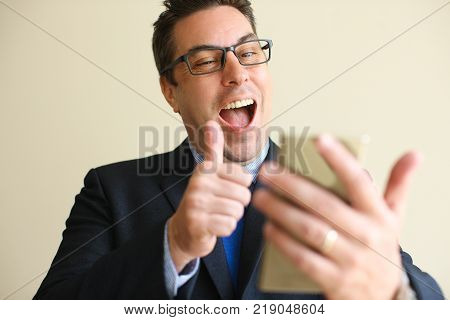 Positive successful businessman showing thumb-up while speaking via video link on smartphone. Cheerful excited man in eyeglasses having online communication. Technology concept
