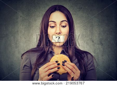 Woman on diet restriction with question mark on her mouth looking at tasty burger isolated on gray background