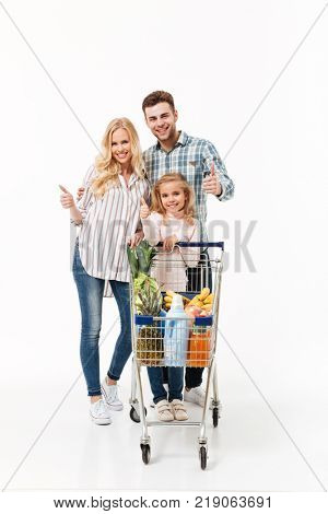 Full length portrait of a happy family standing with a shopping trolley full of groceries and showing thumbs up gesture isolated over white background