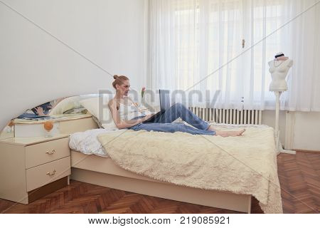 one woman slow culture relaxing laying in bed using laptop computer. ordiray large white room windows daylight.