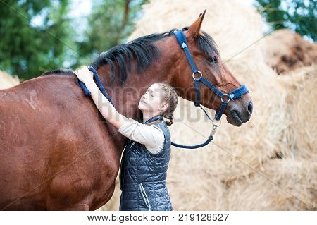 Young cheerful teenage lady equestrian hugging with love her favorite chestnut horse at ranch. Vibrant colored outdoors horizontal summertime image.