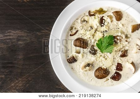 A closeup of a plate of a mushroom cream soup with champignons, cremini, fresh parsley, with grated cheese, on a dark rustic background texture, with a place for text