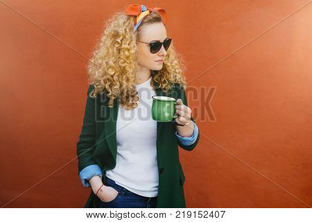 Portrait of pretty woman with curly hair wearing headband stylish sunglasses and green jacket holding green cup of tea in one hand and other hand in pocket looking aside isolated over orange wall