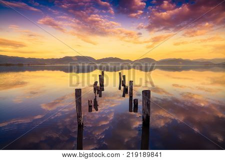 Wooden pier or jetty remains on warm lake sunset and sky reflection water. Versilia Massaciuccoli Tuscany Italy.