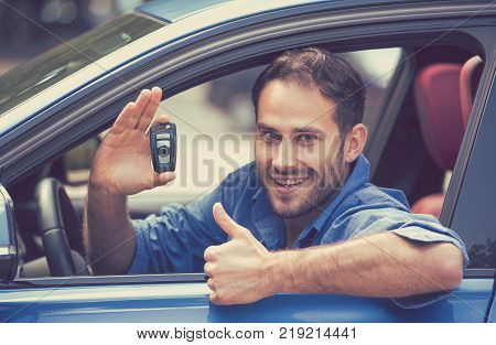 Happy smiling young man sitting in his new blue car showing keys thumbs up isolated outside dealership lot. Personal transportation purchase concept