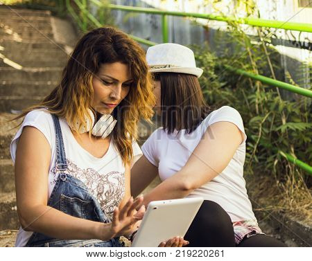 two adult girls sitting in the shade on the stairs listening to music and photographing with a tablet
