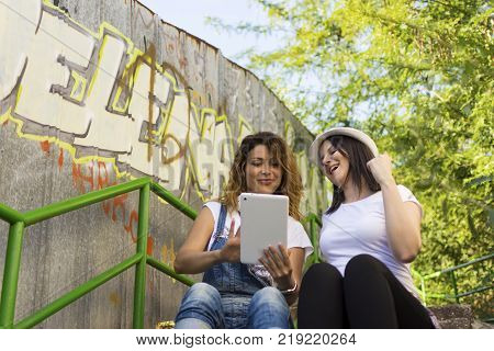 two adult girls sitting in the shade on the stairs listening to music and photographing with a tablet
