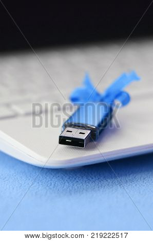Brilliant blue usb flash memory card with a blue bow lies on a blanket of soft and furry light blue fleece fabric beside to a white laptop. Classic female gift design for a memory card