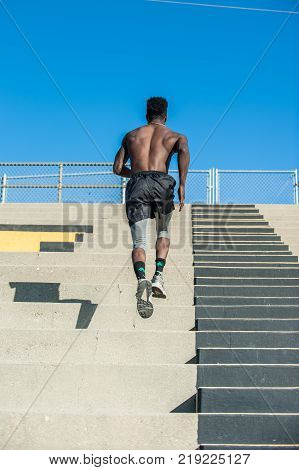 Muscular Jamaican athlete sprinting up plyometric workout at stadium steps in rear view.