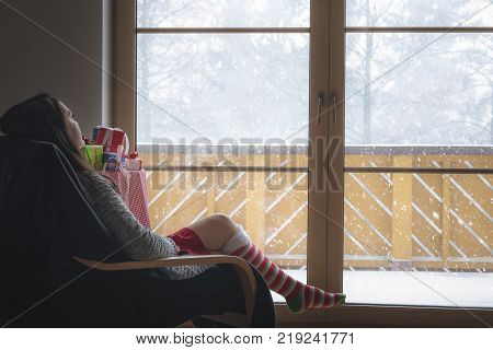 Woman in armchair in front of a snowy window - Young brunette woman wearing a cozy sweater and long red and white socks sitting on an armchair in front of big glass door looking at the snowfall.