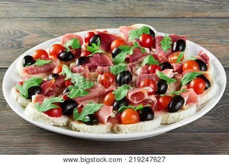 Delicious restaurant appetizer : canapees with white bread, bacon rolls, fresh cherry tomatoes and green parsley leaves. Tasty snack for catering or buffet. A plate with stands on the wooden table