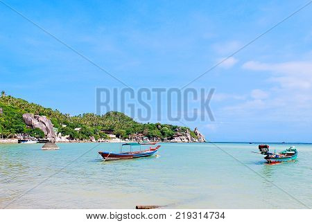 Beautiful seascape in summer season with aqua and blue sea with long tail boats floating on big rock and coconut trees near small port bugalow on coastside white clouds and blue sky.
