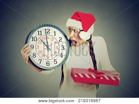 Closeup portrait worried stressed in a hurry young woman wearing red santa claus hat holding clock gift box isolated gray background. Emotion funny face expression last minute christmas shopping