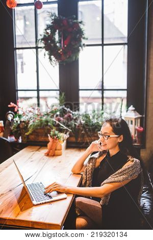 Beautiful Young Brunette Woman Inside Cafe With Christmas Interior At Wooden Table By The Window Tal