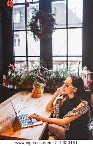 Beautiful Young Brunette Woman Inside Cafe With Christmas Interior At Wooden Table By The Window Tal
