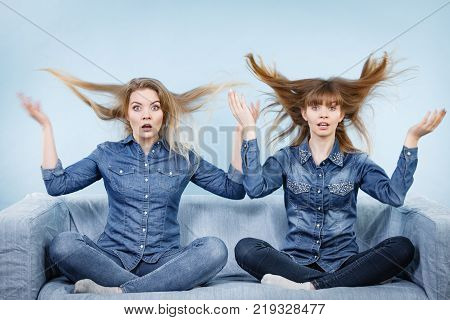 Two shocked women wearing jeans shirts having windblown blonde hair. Blue background.