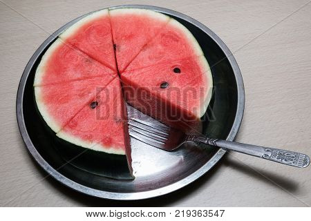 Slices watermelon with fork on steel tray.