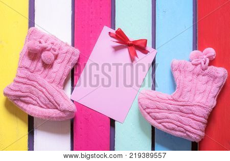 Knitted bootees and a pink paper note - Congratulations on childbirth concept with a message card tied with a red bow surrounded by cute knitted bootees on a colorful wooden background.