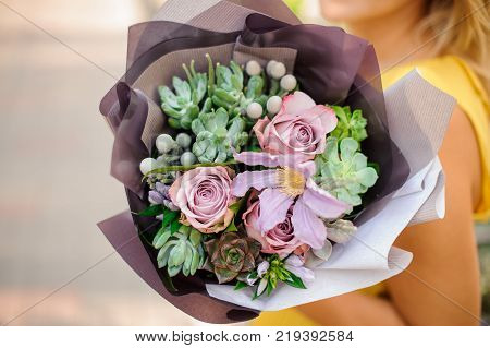 A womans hand is holding a bouquet of flowers including rose flowers echeveria brunia succulents and clematis packed in wrapping-paper