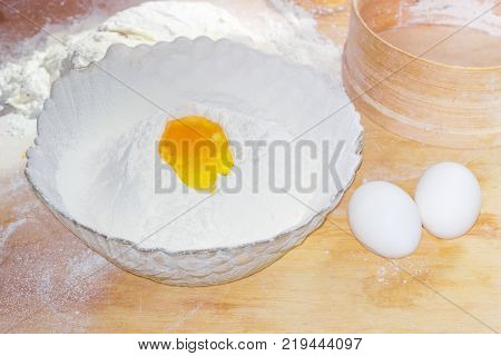 Bowl with sifted wheat flour and broken egg in it and two whole eggs beside on a blurred background of kneaded dough and wooden sieve on the table