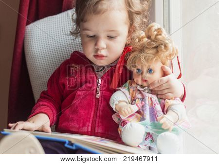 a little girl sitting on the window with her doll and reading a book together
