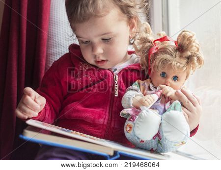 a little girl sitting on the window with her doll and reading a book together