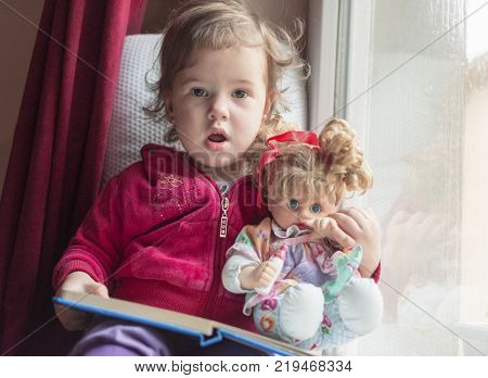 a little girl sitting on the window with her doll and reading a book together