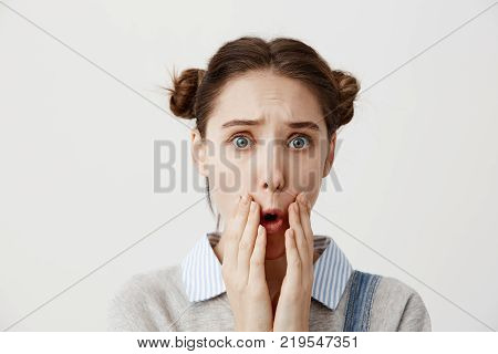Brunette woman being deeply saddened by awful news covering open mouth with hands. Female person with hair in double buns looking on camera in frustration can't believe in grief. Human reactions