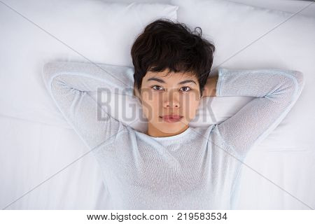 Calm relaxed Asian lady lying with hands behind head and looking at camera. Serious beautiful young woman resting on comfortable bed. Bedtime concept