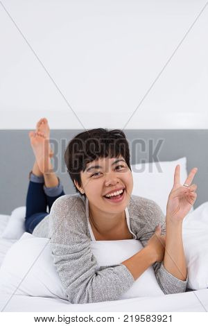 Ecstatic Asian girl showing peace sign lying on bed. Cheerful excited young woman looking at camera and relaxing in bedroom. Joy concept