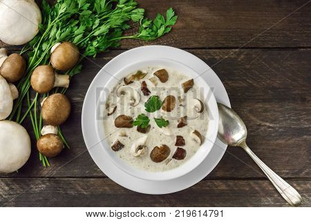 An overhead photo of a plate of a mushroom cream soup with champignons, cremini, and fresh parsley, on a dark rustic background with a spoon and a place for text