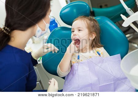 Child at dentist office sits in comfortable chair with wide open mouth and finger in it. Kid shows to doctor in uniform and sterile rubber gloves where she has ache.