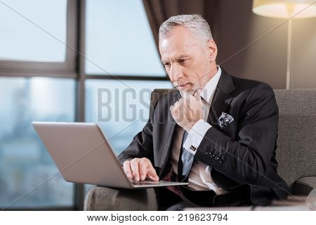 Business prospectives. Wistful senior focussed man staring at the screen while sitting and touching his chin