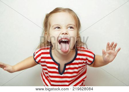 little girl shows her tongue in funny grimace with body leaned toward camera, dressed in striped T-shirt, and stands beside white wall emotional portrait photo.