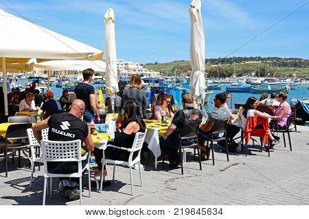 MARSAXLOKK, MALTA - APRIL 1, 2017 - Tourists relaxing at a pavement cafes along the waterfront with traditional Maltese fishing boats in the harbour and views towards the town Marsaxlokk Malta Europe, April 1, 2017.